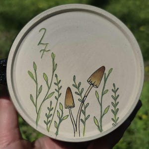 May include: A round, off-white ceramic coaster featuring a hand-drawn design of mushrooms and foliage. The design includes two brown mushrooms with black outlines and green leaves. The number "27" is also visible.
