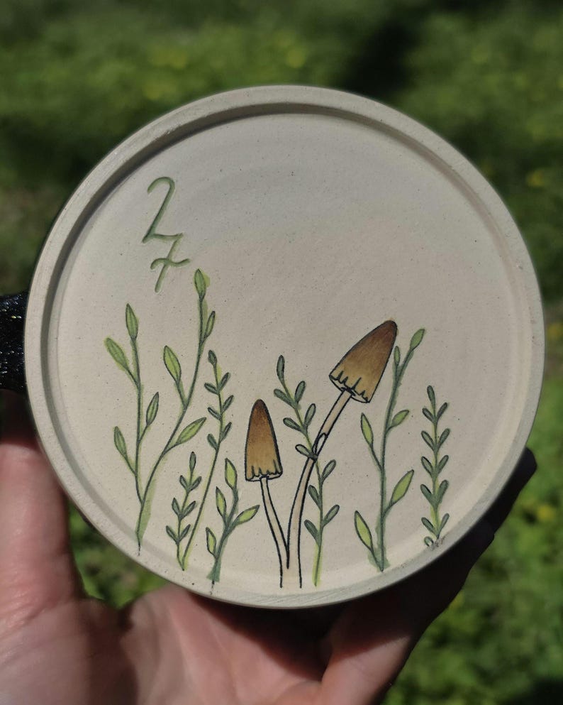 May include: A round, off-white ceramic coaster featuring a hand-drawn design of mushrooms and foliage. The design includes two brown mushrooms with black outlines and green leaves. The number "27" is also visible.