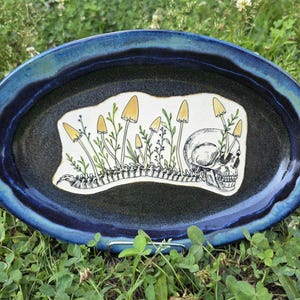 May include: Oval ceramic platter with a dark blue rim and a black center. The center features a white design with a skull, mushrooms, and a spine. The mushrooms have yellow caps and green stems.