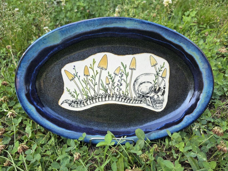 May include: Oval ceramic platter with a dark blue rim and a black center. The center features a white design with a skull, mushrooms, and a spine. The mushrooms have yellow caps and green stems.