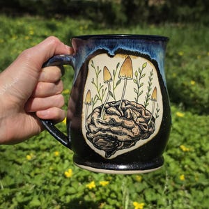 May include: A ceramic mug with a black and blue speckled exterior. The mug features a detailed illustration of a brain with mushrooms and foliage. The handle is black, and the mug is held against a green background.