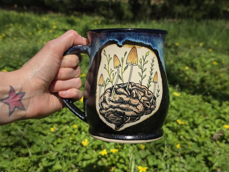 May include: A ceramic mug with a black and blue speckled exterior. The mug features a detailed illustration of a brain with mushrooms and foliage. The handle is black, and the mug is held against a green background.
