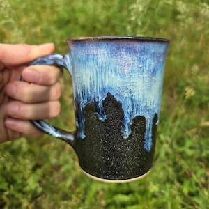 May include: A handmade ceramic mug with a blue and black glaze. The mug has a handle and a unique drip effect, with the blue glaze cascading down the black base. The mug is held in a hand against a blurred green background.