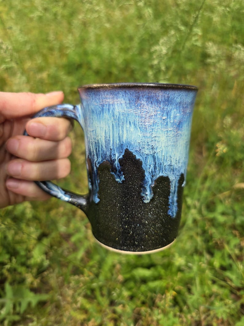 May include: A handmade ceramic mug with a blue and black glaze. The mug has a handle and a unique drip effect, with the blue glaze cascading down the black base. The mug is held in a hand against a blurred green background.