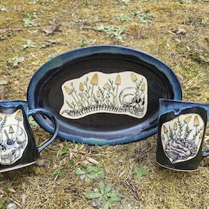 May include: A ceramic set featuring a dark blue oval platter and two mugs. The platter and mugs have a black interior with a blue rim. Each piece has a unique illustration of mushrooms, a skull, and a spine.