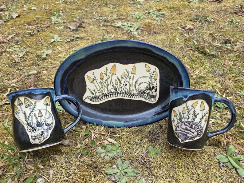 May include: A ceramic set featuring a dark blue oval platter and two mugs. The platter and mugs have a black interior with a blue rim. Each piece has a unique illustration of mushrooms, a skull, and a spine.