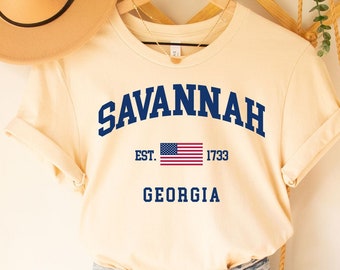 Puede incluir: Una camiseta de manga corta color crema con texto azul que dice SAVANNAH, EST. 1733 y GEORGIA, con una pequeña bandera estadounidense. La camiseta se combina con pantalones cortos de mezclilla azul claro desgastados, un fedora beige y un bolso de macramé.