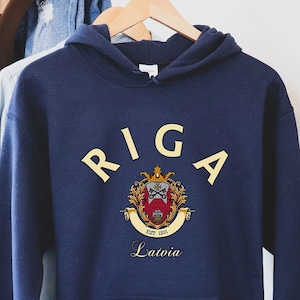 Puede incluir: Una sudadera con capucha azul marino con un estampado gráfico blanco del escudo de la ciudad de Riga y el texto "RIGA" en amarillo. El escudo incluye un escudo rojo con una cruz blanca y una corona dorada. El texto "Letonia" está impreso debajo del escudo.