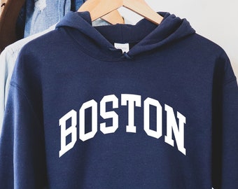 Puede incluir: Una sudadera con capucha azul marino con la palabra "BOSTON" impresa en blanco en la parte delantera. La sudadera está colgada de una percha.
