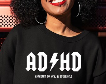 May include: Black crewneck sweatshirt with the text "ADHD" in a white, bold, rock-and-roll style font, with the phrase "Highway to Hey, A Squirrel!" below. The sweatshirt is worn by a person with dark curly hair and hoop earrings.