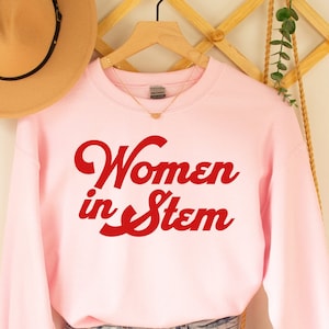 May include: Pink sweatshirt with the text "Women in Stem" in red lettering. The sweatshirt is hanging on a wooden rack with a brown hat and a woven bag.