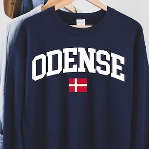 Puede incluir: Una sudadera azul marino de cuello redondo con el texto blanco ODENSE en una fuente universitaria arqueada sobre una pequeña bandera danesa rectangular. La prenda de manga larga se muestra colgada en una percha de madera.