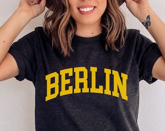 May include: A dark grey t-shirt with the word "BERLIN" printed in yellow on the front. The shirt is being worn by a person with brown hair and a brown hat.