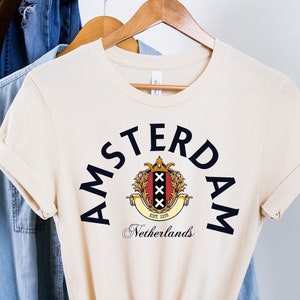 Może przedstawiać: Kremowa koszulka z czarnym nadrukiem herbu Amsterdamu i tekstem "Amsterdam Holandia".