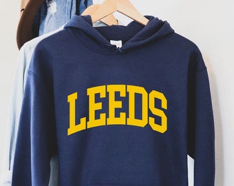 Puede incluir: Sudadera con capucha azul marino con la palabra "LEEDS" en letras amarillas. La sudadera tiene un bolsillo delantero y una capucha. Otras prendas de vestir y un sombrero cuelgan en un perchero en el fondo.