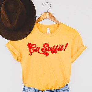 May include: Yellow t-shirt with red text that says "Ca Suffit!"  The shirt is on a hanger with a brown hat on top.  The shirt is paired with denim shorts.