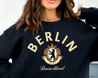 May include: Black sweatshirt with a gold graphic of the Berlin coat of arms. The graphic includes the text "BERLIN" in a gold script font, a bear, a crown, and the text "EST. 1237" and "Deutschland".