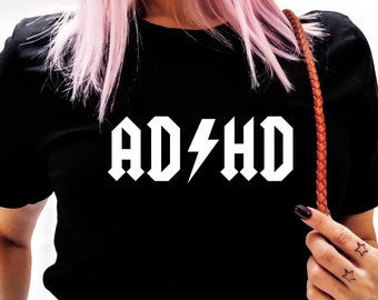 Puede incluir: Camiseta negra con texto blanco que dice "AD/HD" en una fuente de rock and roll con un rayo en lugar de la barra oblicua.