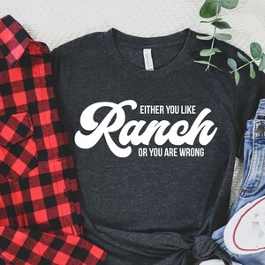 May include: A dark grey t-shirt with white text that reads "Either you like Ranch or you are wrong". The text is in a retro font style. The shirt is laid out on a white surface with a red and black plaid shirt, blue jeans, and white sneakers.