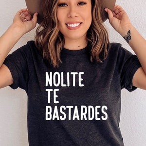 May include: Dark gray t-shirt with the text "NOLITE TE BASTARDES" in white. The person in the image is wearing a brown hat and blue jeans. The shirt is a casual style.