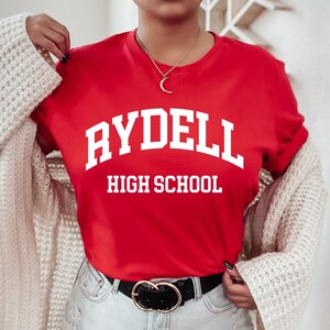 Può includere: Una maglietta rossa con testo bianco che dice "Rydell High School".