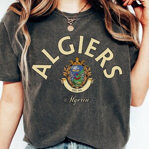Peut inclure: Un t-shirt gris anthracite à manches courtes avec le texte ALGIERS en grandes lettres jaunes arquées au-dessus d'un emblème coloré. Sous l'emblème, le mot Algeria est imprimé en écriture noire avec le texte EST. 944 sur une bannière.