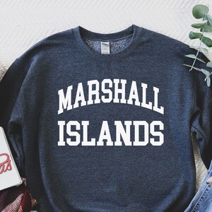 Puede incluir: Una sudadera azul oscuro con el texto "MARSHALL ISLANDS" en letras blancas. La sudadera está colocada sobre una superficie blanca con una bufanda de cuadros, un cartel que dice "Feliz Navidad" y un par de vaqueros y botas.