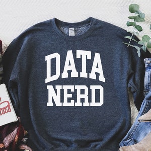 May include: A dark blue sweatshirt with the text "DATA NERD" in white letters. The sweatshirt is laid flat on a white surface with a plaid scarf and a pair of jeans and boots.