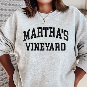 May include: A light gray sweatshirt with the words "MARTHA'S VINEYARD" printed in bold, black, arched letters. The sweatshirt has long sleeves and a crew neck.