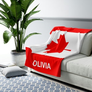 May include: A red and white Canadian flag blanket with the name Olivia printed on the bottom. The blanket is draped over a white couch.