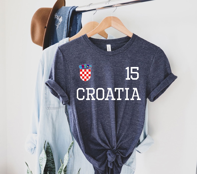May include: Navy blue t-shirt with the word "CROATIA" in white block letters. The number "15" is above the text, and a Croatian coat of arms is on the upper left. The shirt is hanging on a wooden hanger.