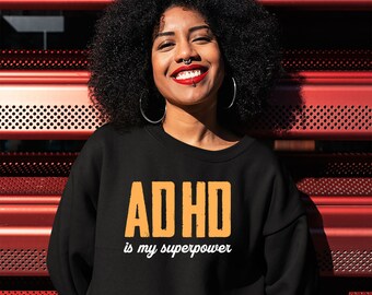 May include: Black sweatshirt with the text "ADHD is my superpower" in orange and white. The person in the image is smiling and wearing hoop earrings and a lip piercing. The background is a red metal structure.