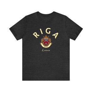 May include: A dark grey t-shirt with a cream colored graphic of the coat of arms of Riga, Latvia. The graphic includes the text "RIGA" in a cream colored arch above the coat of arms and the text "EST. 1201" and "Latvia" below the coat of arms.