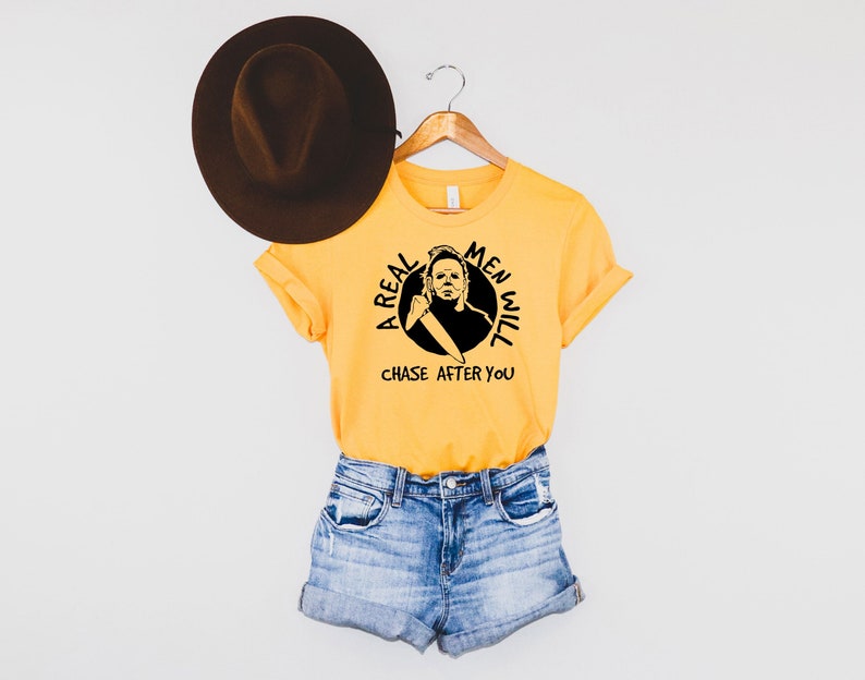 May include: A yellow t-shirt featuring a black and white graphic of a man holding a knife. The graphic is in a circle with the text "A Real Men Will Chase After You".