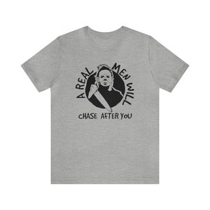 May include: A grey t-shirt with a black graphic of a man holding a knife. The graphic is in a circle with the text "A Real Men Will Chase After You".