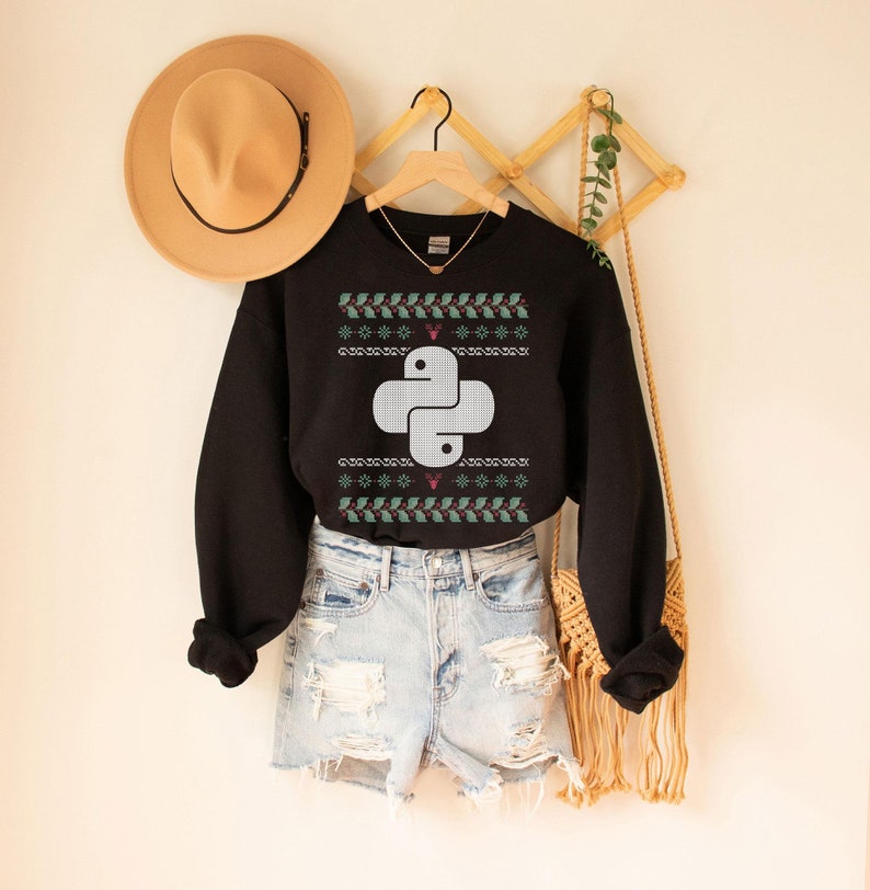 Puede incluir: Sudadera negra con un logotipo de Python blanco sobre un patr&oacute;n de su&eacute;ter navide&ntilde;o verde y rojo. La sudadera se lleva sobre unos pantalones cortos de mezclilla azul claro.