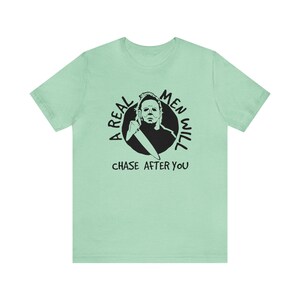 May include: A light green t-shirt featuring a black and white graphic of a man holding a knife. The graphic is in a circle with the text "A Real Men Will Chase After You".