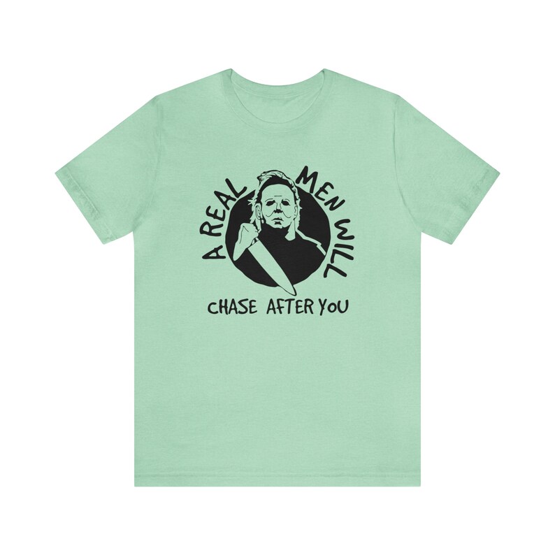 May include: A light green t-shirt featuring a black and white graphic of a man holding a knife. The graphic is in a circle with the text "A Real Men Will Chase After You".