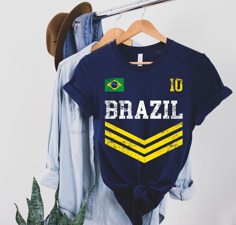 May include: A navy blue t-shirt with a white distressed graphic of the word "BRAZIL" and the number "10". The shirt also features a small yellow and green Brazilian flag and two yellow chevron stripes.