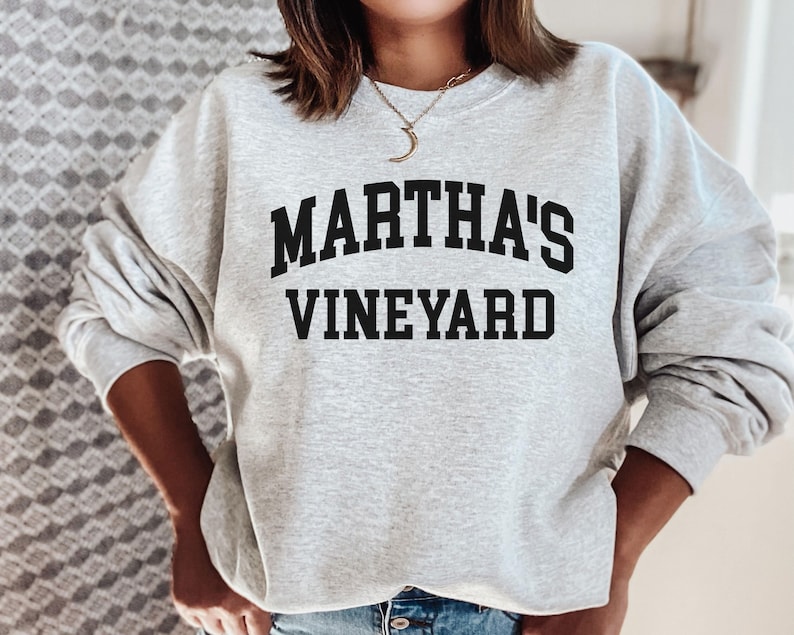 May include: A light gray sweatshirt with the words "MARTHA'S VINEYARD" printed in bold, black, arched letters. The sweatshirt has long sleeves and a crew neck.