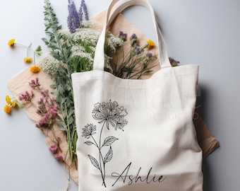 Puede incluir: Una bolsa de lona blanca con un dibujo de línea negra de una flor y el nombre "Ashlie" impreso en la parte delantera.