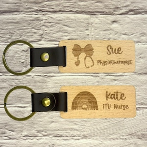 May include: Two wooden keychains with leather straps and metal rings. One keychain has the name "Sue" and the text "Physiotherapist" with a bow and stethoscope design. The other keychain has the name "Kate" and the text "ITU Nurse" with a rainbow design.