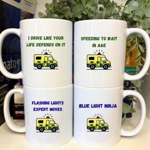 May include: Four white ceramic mugs with black handles, each printed with a different phrase and an ambulance graphic. The phrases read: "I drive like your life depends on it", "Speeding to wait in A&E", "Flashing lights expert moves", and "Blue light ninja."