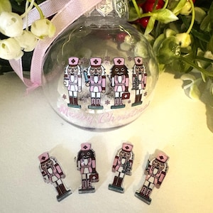 May include: A clear glass Christmas ornament with pink and white nutcracker figures and the text "Merry Christmas." The ornament has a pink ribbon for hanging. Several nutcracker figures are scattered below.
