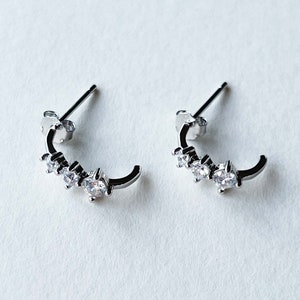 Könnte beinhalten: Ein Paar silberne Creolen mit kleinen, klaren Zirkonia-Steinen, die in das Metall gefasst sind. Die Ohrringe sind in einem schlichten und eleganten Design gehalten.