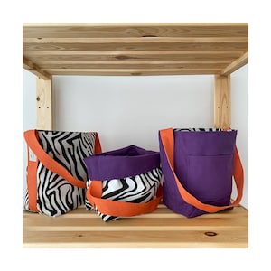 Puede incluir: Tres bolsas de mano con asas naranjas. Dos bolsas son de estampado de cebra en blanco y negro y una es morada con un bolsillo. Las bolsas est&aacute;n en un estante de madera.