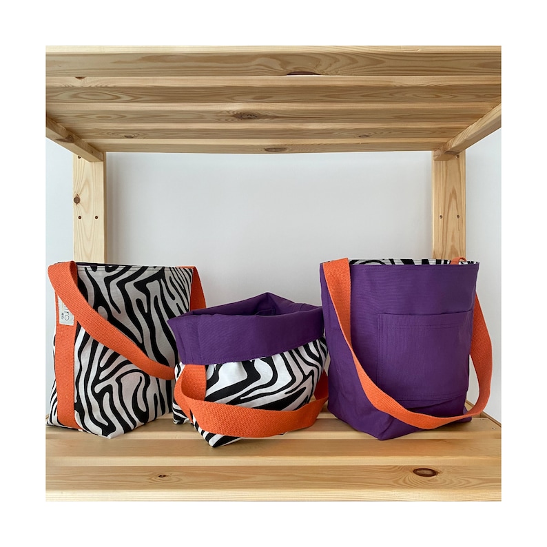 Puede incluir: Tres bolsas de mano con asas naranjas. Dos bolsas son de estampado de cebra en blanco y negro y una es morada con un bolsillo. Las bolsas est&aacute;n en un estante de madera.