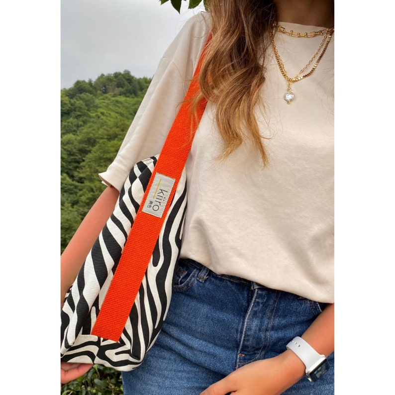 Puede incluir: Una bolsa de mano de lona con estampado de cebra en blanco y negro con una correa naranja. La bolsa tiene una peque&ntilde;a etiqueta blanca con la palabra "Kiliro" impresa en ella.