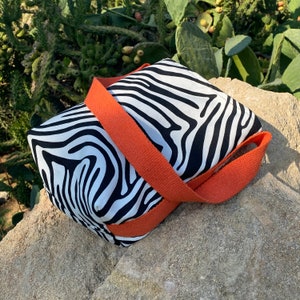Puede incluir: Una bolsa de mano de tela con estampado de cebra en blanco y negro con una correa naranja. La bolsa est&aacute; hecha de tela y tiene un cierre de cremallera.