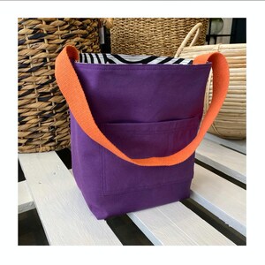 Puede incluir: Bolso tote morado con correas naranjas. El bolso tiene un peque&ntilde;o bolsillo en la parte delantera y est&aacute; hecho de un tejido resistente.